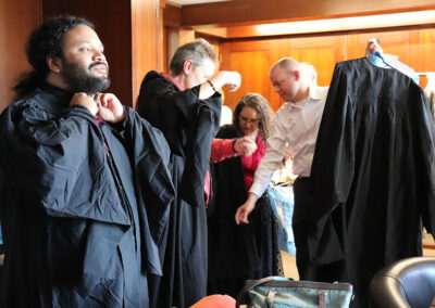 Four people are in a room getting dressed in black graduation gowns. Two of them are putting the gowns on over their clothes, while a third person helps another adjust their gown. The room, with its wooden walls and counter in the background, buzzes with the anticipation of an important event.