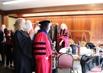 A group of people in black and red graduation robes are preparing for a ceremony in the wood-paneled room of the Unitarian Universalist Church of Berkeley. Some are helping each other with the robes, while others are gathered around a table with various items on it, making final event preparations.