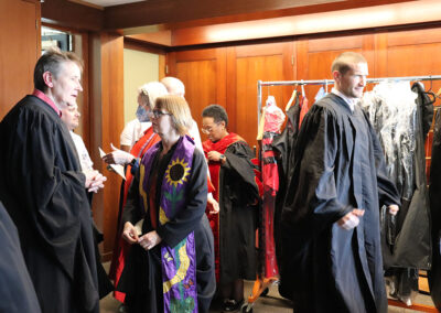 A group of people in academic regalia are gathered in a room, some chatting while others put on their robes. In the background, a clothing rack with additional garments is visible. The atmosphere is one of preparation and anticipation at the Unitarian Universalist Church of Berkeley.