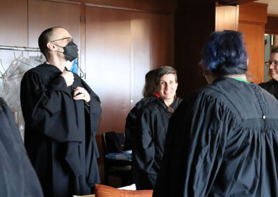 A group of people wearing black robes are gathered in a room, engaged in lively conversation. One person on the left adjusted their mask while others smiled and chatted. The wooden walls and an empty clothing rack set the backdrop.