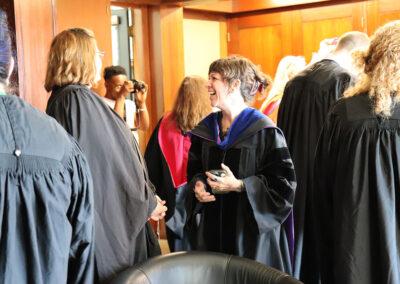 A group of people in academic regalia gather in a warmly lit room. Two people in black robes with colorful hoods stand in the foreground, smiling and chatting. Other figures are conversing in the background, while one person takes photos, capturing the joyous moments of this event.