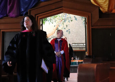 A group of people dressed in academic regalia walk down an aisle. Multicolored fabric hangs from the ceiling above them. A sign reading "Black Lives Matter" is visible in the background, adding a powerful message to the formal and ceremonial atmosphere.