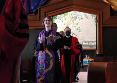 A procession of faculty members in colorful robes walks down the aisle of a church. One person in the foreground wears a purple robe with a multicolored stole. The church, part of the Unitarian Universalist Church of Berkeley, is adorned with draped, multicolored fabric. A "Black Lives Matter" sign is visible in the background.