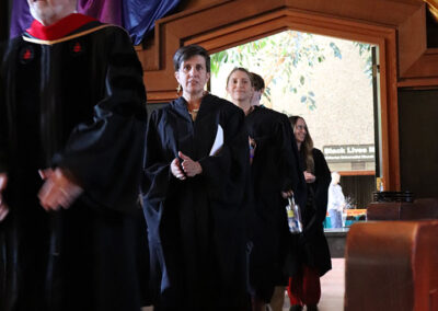 A group of people in academic robes walk in a procession at the Unitarian Universalist Church of Berkeley. They are indoors, with colorful drapes hanging from the ceiling and a lit sign that reads "Black Lives" visible in the background.