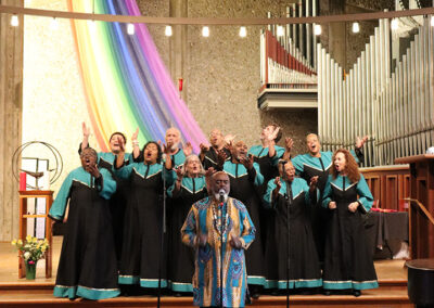 At the Unitarian Universalist Church of Berkeley, a choir dressed in black robes with teal accents sang joyfully in a church. A conductor wearing a colorful robe led the harmonious voices, singing into a microphone. Behind them, a large pipe organ stood majestically as rainbow fabric draped the wall.