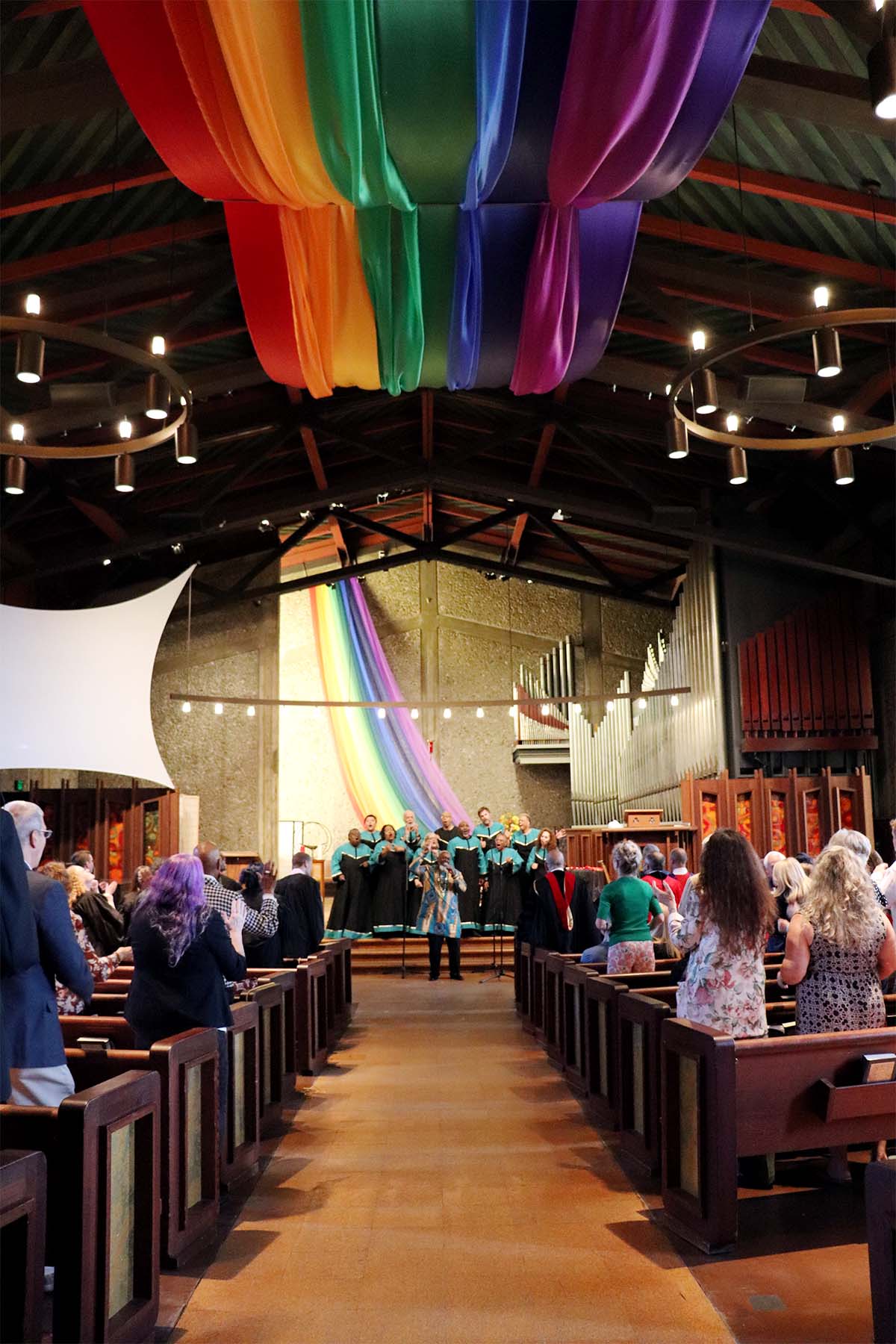 The Unitarian Universalist Church of Berkeley hosted the 2024 Commencement ceremony where a choir performed on a stage at the front of the church, adorned with large, vibrant rainbow banners hanging from the ceiling. The audience in the pews watched attentively.