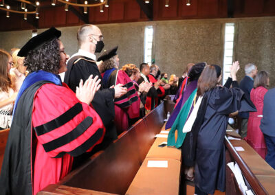 A group of people in academic regalia, including caps and gowns, are sitting in pews at an indoor ceremony. They are clapping and dancing while celebrating. Some attendees are standing while others remain seated.