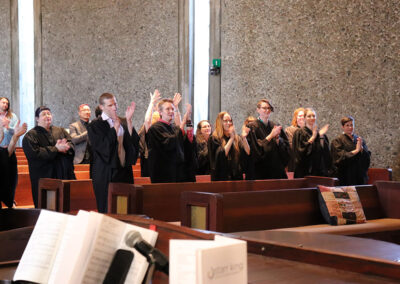 A group of people dressed in black graduation robes stand and applaud in a church setting during Commencement. Behind them are wooden pews and a stone wall. In the foreground, there is a microphone and an open book of sheet music.