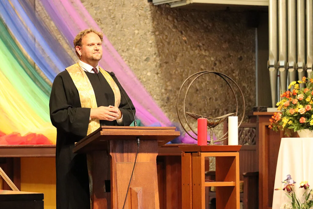 A person stands at a wooden podium, wearing a black robe with a gold stole. Behind them, colorful, flowing fabric in rainbow hues drapes across a stone wall. On the right is a floral arrangement and a small table with a chalice, as well as a red and white candle.