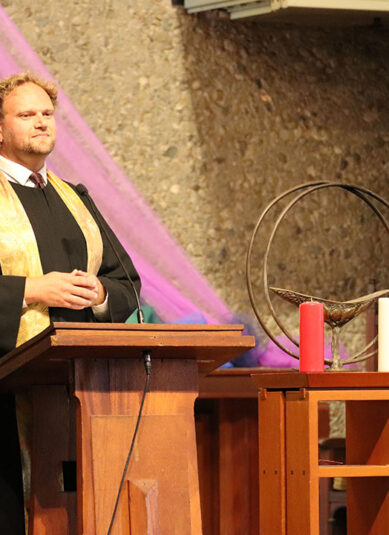 A person stands at a wooden podium, wearing a black robe with a gold stole. Behind them, colorful, flowing fabric in rainbow hues drapes across a stone wall. On the right is a floral arrangement and a small table with a chalice, as well as a red and white candle.