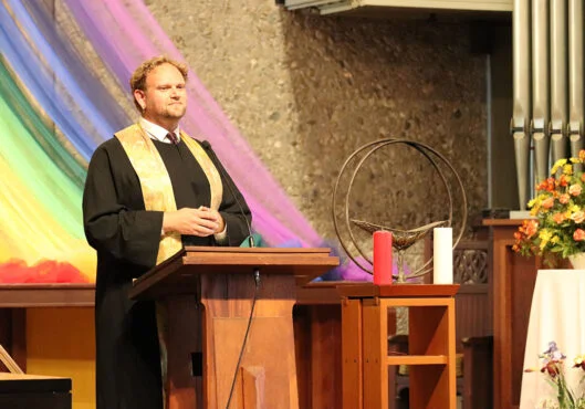 A person stands at a wooden podium, wearing a black robe with a gold stole. Behind them, colorful, flowing fabric in rainbow hues drapes across a stone wall. On the right is a floral arrangement and a small table with a chalice, as well as a red and white candle.