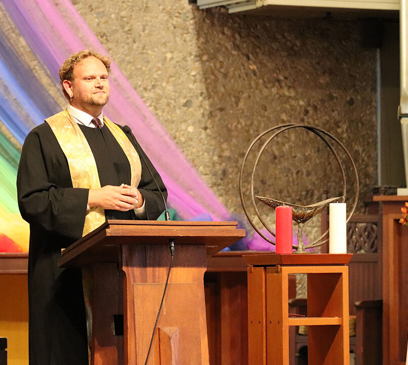 A person stands at a wooden podium, wearing a black robe with a gold stole. Behind them, colorful, flowing fabric in rainbow hues drapes across a stone wall. On the right is a floral arrangement and a small table with a chalice, as well as a red and white candle.