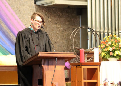A person with short hair and glasses, wearing a black robe, stands at a wooden lectern, speaking into a microphone. Behind them is a decorative background with rainbow fabric, a metal chalice, flowers, and organ pipes.