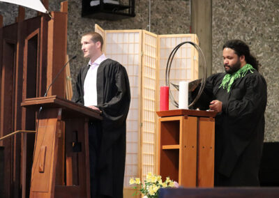 Two individuals dressed in black academic robes are positioned indoors. One speaks at a lecturn, while the other stands beside a table with a chalice, a red candle, and a white candle. A screen and flowers enhance the formal setting.