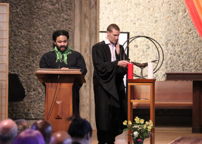 Two people stand at the front of a room. The person on the left is speaking at a lecturn, wearing black robes with green detailing. The person on the right, also in black robes, is adjusting a candle on a stand. In the foreground, members of the Unitarian Universalist Church of Berkeley are seated and watching this event.