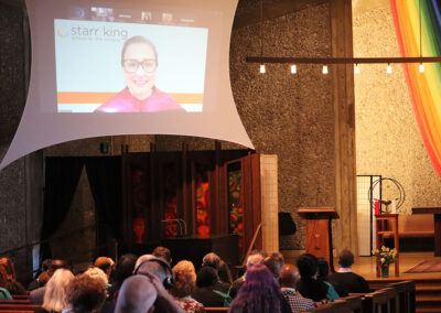 An audience sits in a chapel facing a large screen displaying a video conference with a speaker identified by the text "Starr King School for the Ministry." A rainbow banner decorates the right side of the chapel, marking an inclusive gathering.
