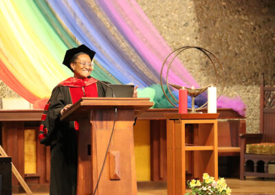A person wearing academic regalia, including a cap and red and black robe, stands at a wooden lectern and speaks into a microphone. Behind them, rainbow-colored fabric drapes against a textured wall while a chalice, red candle, and white candles adorn the stage area.