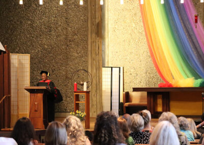 A person in academic regalia speaks at a podium on a stage decorated with a large, colorful rainbow banner. Seated attendees at the Unitarian Universalist Church of Berkeley watch the speaker. The backdrop features a textured wall with vertical light fixtures hanging from the ceiling.