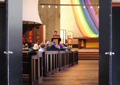 A church interior with open double doors reveals an ongoing event at the Unitarian Universalist Church of Berkeley. Attendees are seated in wooden pews facing a speaker at a lectern. The altar is adorned with a large, colorful rainbow fabric hanging from the ceiling, and the floor is tiled in a reddish-brown hue.