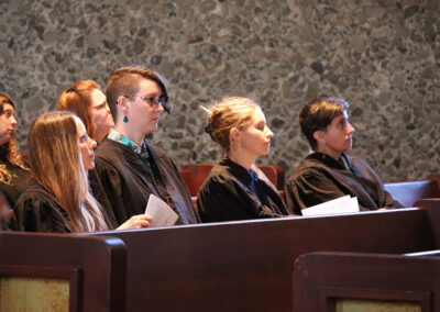 A group of people are sitting in wooden pews, wearing black robes and holding programs. They appear attentive and focused as they engage in the ceremony. The background features a stone wall.