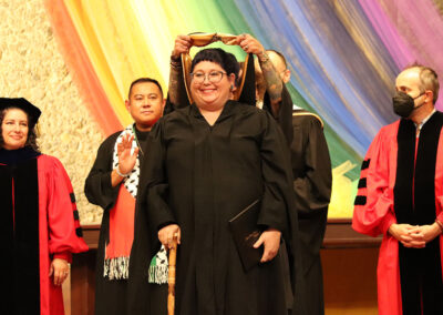 A beaming graduate in a black gown is being hooded on stage by a faculty member during the Commencement ceremony at the Unitarian Universalist Church of Berkeley. Faculty members in red and black academic regalia stand around them, with a colorful rainbow backdrop behind them creating an unforgettable event.
