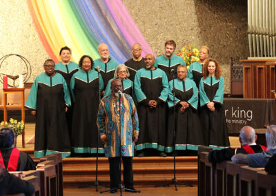 A choir in green and black robes stands on stage in front of a colorful rainbow cloth backdrop. A person in a vibrant, patterned shirt speaks into a microphone. People sit in pews facing the choir, with an organ visible on the right.