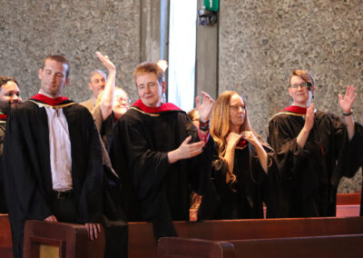 A group of graduates in black caps and gowns with red accents dance and clap inside a building. The background features rough-textured walls. The mood is celebratory.