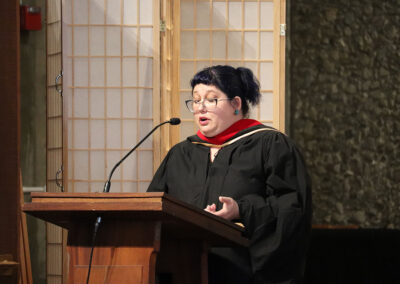 A person wearing academic regalia speaks at a wooden podium with a microphone during the Commencement. The speaker has short, dark hair tied back, round glasses, and is gesturing with one hand. Behind them is a room divider and a textured wall.