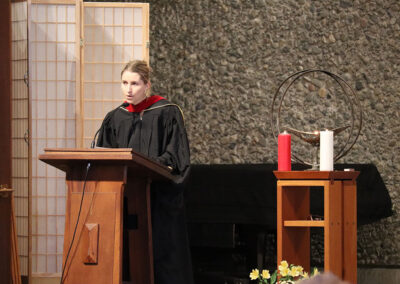 A person in a black graduation gown with a red collar stands at a wooden podium, addressing the audience. The setting includes a large stone wall, a room divider, and a small table with a chalice, two large candles—one red and one white—and some yellow flowers.