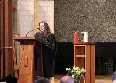 A person wearing academic regalia stands at a wooden podium, speaking into a microphone. Behind them, there is a screen and a table with a chalice, two candles - one red and one white, and a small flower arrangement.