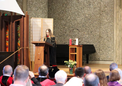 A person in academic regalia stands at a lectern, delivering a speech at the Unitarian Universalist Church of Berkeley. They are surrounded by an audience, some of whom are also in academic attire. The background features a screen divider, a table with a chalice, red and white candles, and a bouquet of flowers.