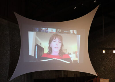 A person appears on a large screen, participating in Commencement virtually. The screen is draped in white, curved fabric. Several other participant windows are visible along the top of the screen.