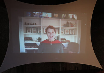 A woman is speaking during Commencement displayed on a large screen. She is wearing a red and black outfit and smiling. Behind her is a white shelf with various decorations and framed pictures. Smaller video thumbnails of other participants are visible at the top.
