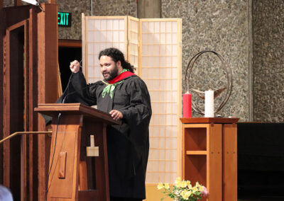 A person wearing academic robes and a green hood stands at a podium, speaking passionately with a raised fist. Behind them is a decorative screen and a ceremonial table with a chalice and red and white candles, along with a metal circular chalice.