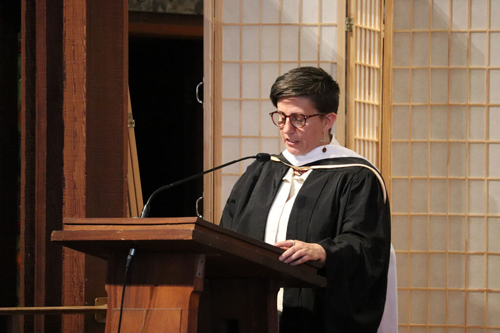 A person wearing a black academic gown and white stole stands at a wooden podium. They have short hair and glasses. The background features wooden panels with lattice designs, providing an elegant backdrop for this memorable event.