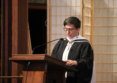 A person wearing a black academic gown and white stole stands at a wooden podium. They have short hair and glasses. The background features wooden panels with lattice designs, providing an elegant backdrop for this memorable event.