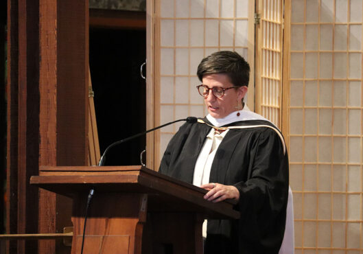 A person wearing a black academic gown and white stole stands at a wooden podium. They have short hair and glasses. The background features wooden panels with lattice designs, providing an elegant backdrop for this memorable event.