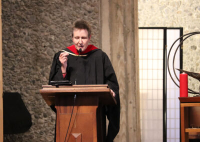 A person wearing academic robes and a red hood speaks into a microphone at a wooden podium. The backdrop includes a textured stone wall and shoji screen panels, with a large candle and metal chalice on a table nearby.