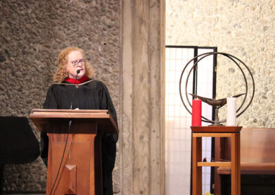 A person with curly hair speaks at a wooden podium inside a stone-walled room. Next to the podium is a stand with a metal chalice and two candles, one red and one white.