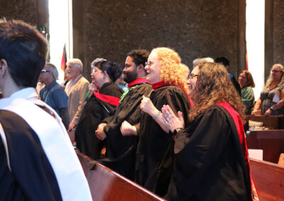 A group of individuals in graduation gowns are standing and clapping in a pew inside the Unitarian Universalist Church of Berkeley, enjoying the moment. They are surrounded by others who are similarly dressed, and light streams in through the tall windows.
