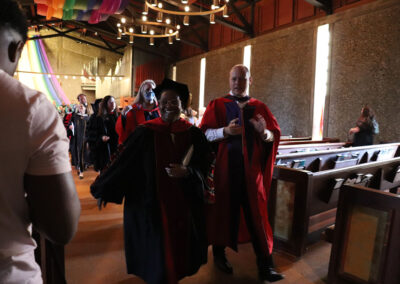 A procession of people wearing academic regalia is walking down the aisle of the sanctuary at the Unitarian Universalist Church of Berkeley. The participants are smiling and appear to be celebrating. The chapel, softly lit with chandeliers, is adorned with ribbon decorations hanging from the ceiling.