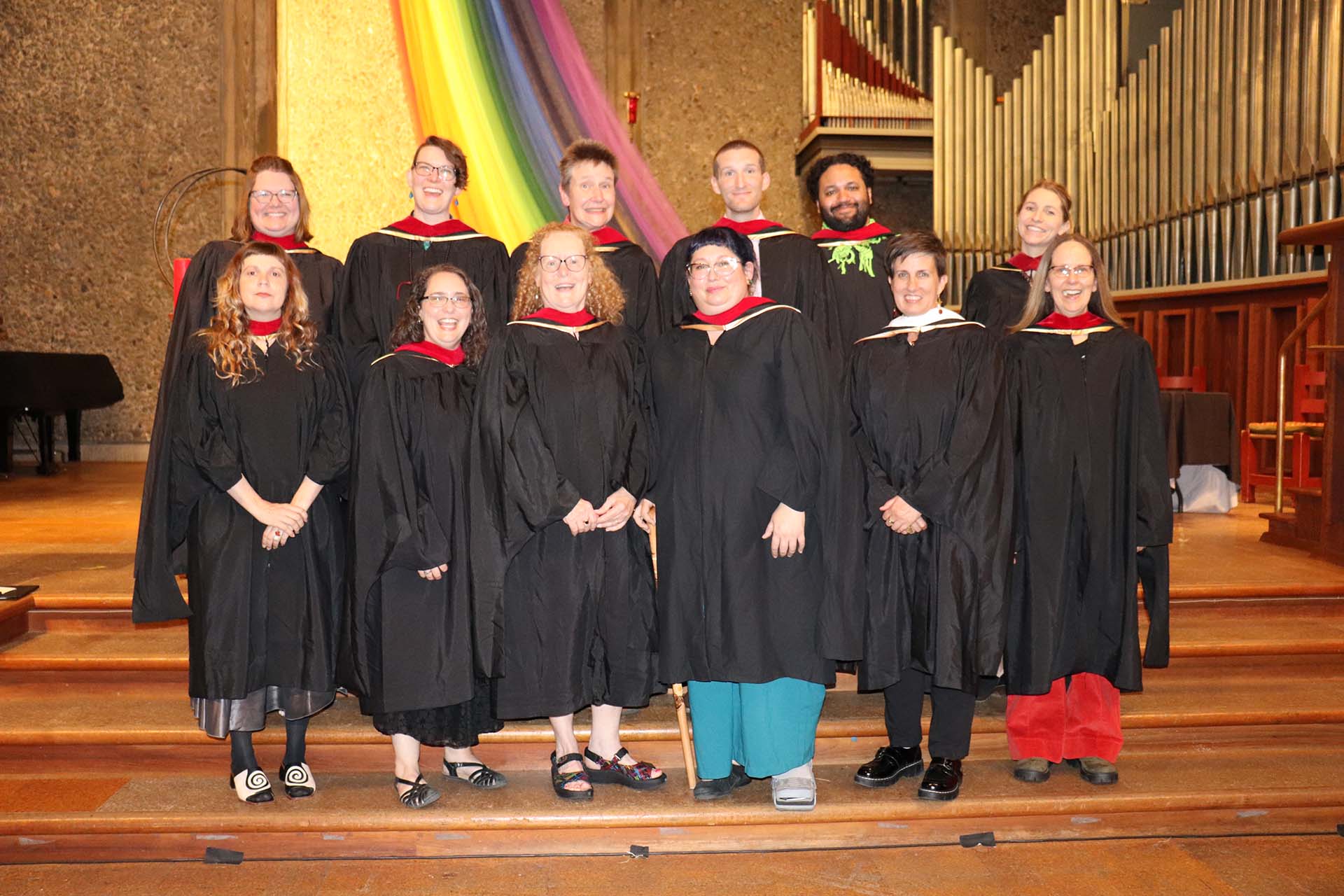 A group of twelve individuals, dressed in academic gowns, stands on steps at the 2024 Starr King Commencement. A large rainbow flag is draped in the background alongside organ pipes. Some people are smiling, and all are looking at the camera.