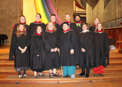 Eleven people wearing graduation robes stand together on a wooden stage in front of a rainbow banner and organ pipes, celebrating their accomplishment.