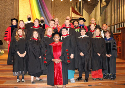 A diverse group of people stands on steps in academic regalia, smiling for the camera at Commencement. Behind them is a colorful fabric drape and a large organ.