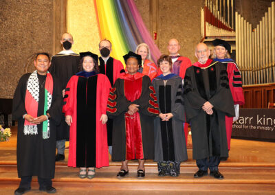 A group of ten formally dressed people stand on stage in academic regalia, posing in front of an organ and a multicolored rainbow backdrop. Some individuals smile.