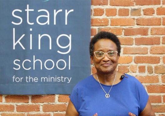 A person wearing glasses and a blue shirt stands in front of a brick wall next to a sign that reads "Starr King School for the Ministry." A microphone is clipped to the shirt, indicating they might be giving a special announcement.