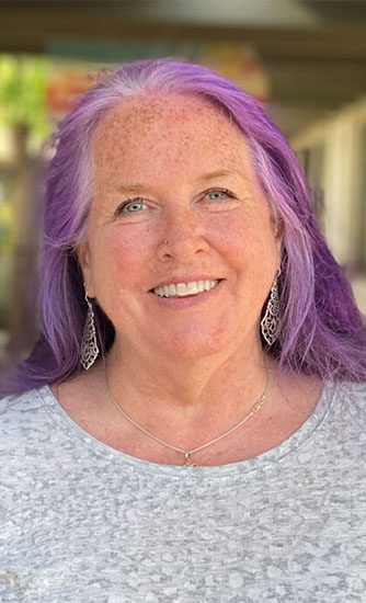 A woman with long, straight purple hair smiles at the camera. She is wearing a light gray textured top, dangling silver earrings, and a delicate necklace. The background is softly blurred, hinting at an outdoor setting with indistinct details.