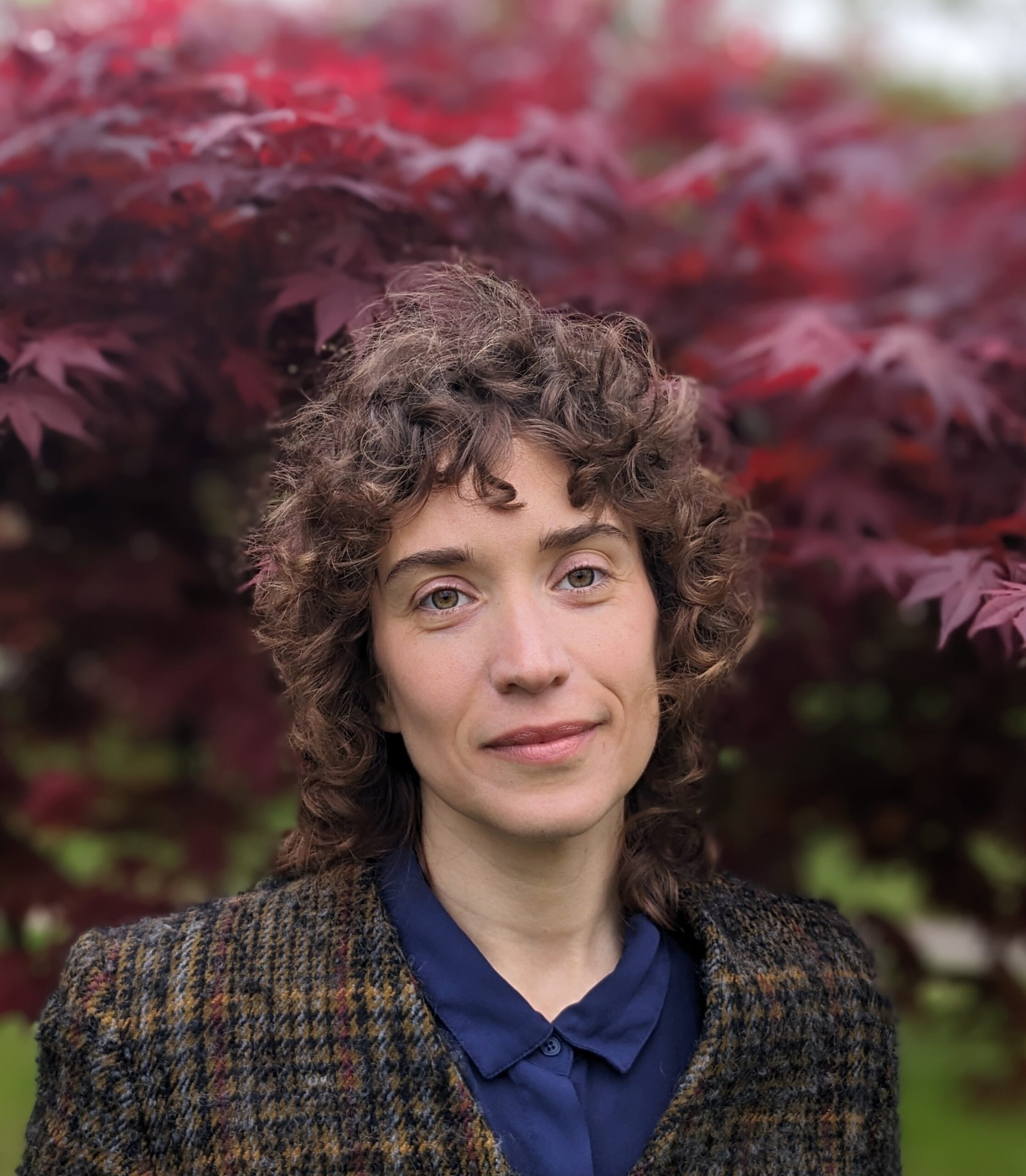 A calm Rabbi with curly hair stands in front of a tree with vibrant red leaves. Wearing a checked coat over a blue shirt, Ora Nitkin-Kaner commands attention as the background softly blurs around them.