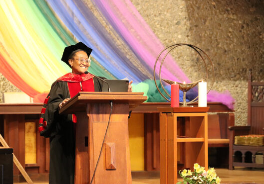 A speaker in academic regalia, including a black cap and red gown, stands at a podium while giving a speech. Behind them is a colorful rainbow fabric draped as a backdrop.
