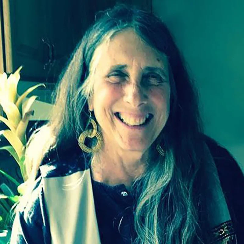 Image of an Arab Jewish woman with jewelry and a gray and black striped top set in front of a dark wooden background